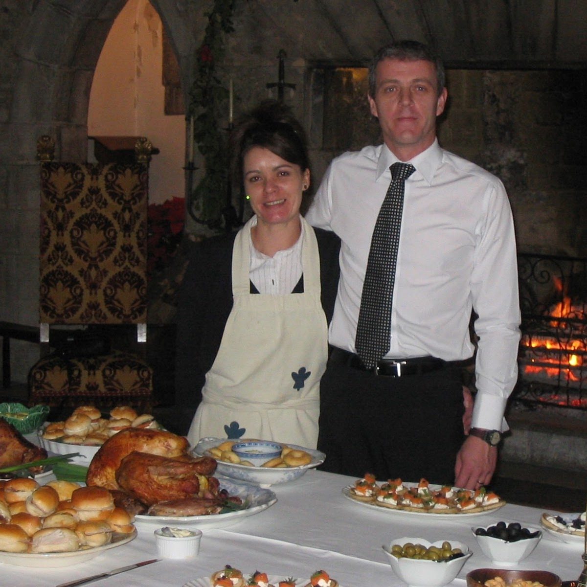 A domestic couple standing in front of a lit fireplace.